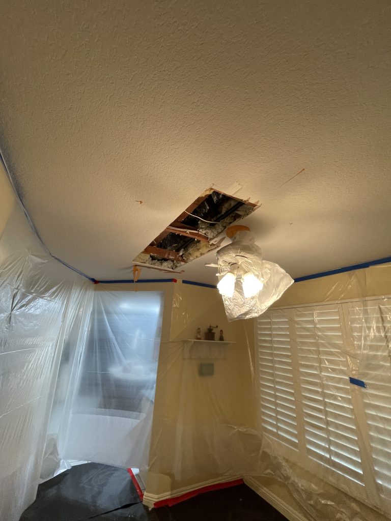 Ceiling drywall cut open to expose framing during a water leak repair, with plastic containment protecting the room below